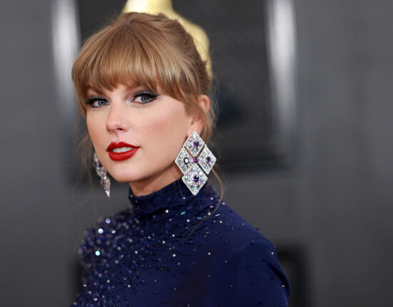 Taylor Swift is looking at the viewer.Her hair is up and she is wearing huge diamond and purple gem earrings. with a navy blue outfit is covered in glittering beads.