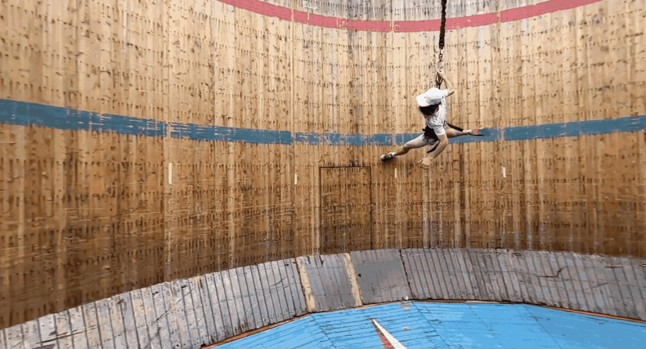 In this gif, a man in a white shirt and hat is running along the interior wall of a wooden cylinder in an attempt to emulate low-gravity exercise on the moon.