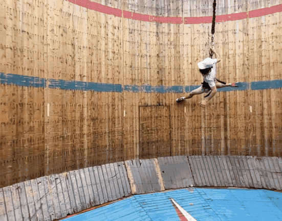 In this gif, a man in a white shirt and hat is running along the interior wall of a wooden cylinder in an attempt to emulate low-gravity exercise on the moon.