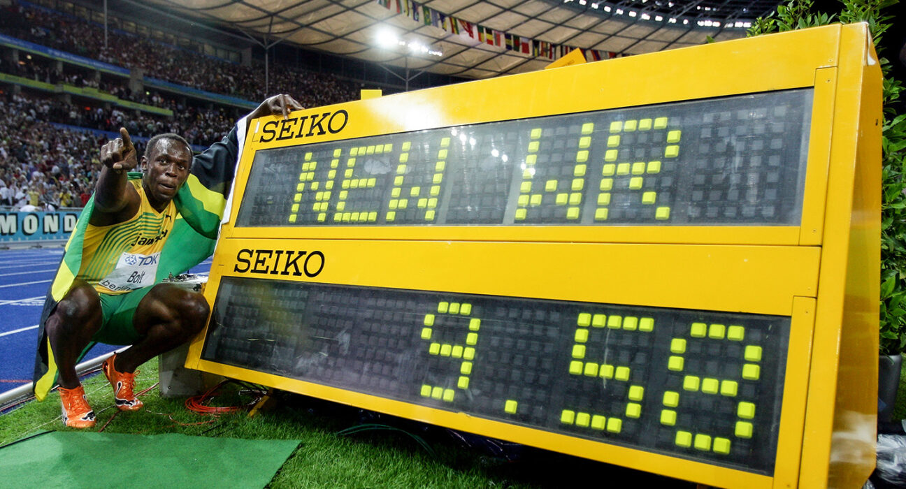 Usain Bolt crouches next to a digital display showing his new world record while pointing to a crowd in a stadium