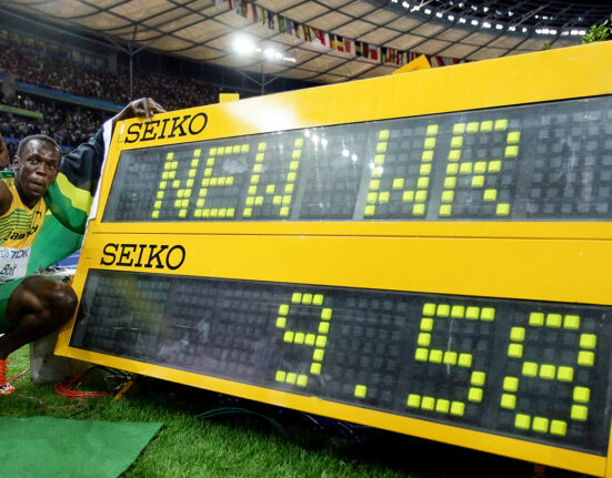 Usain Bolt crouches next to a digital display showing his new world record while pointing to a crowd in a stadium