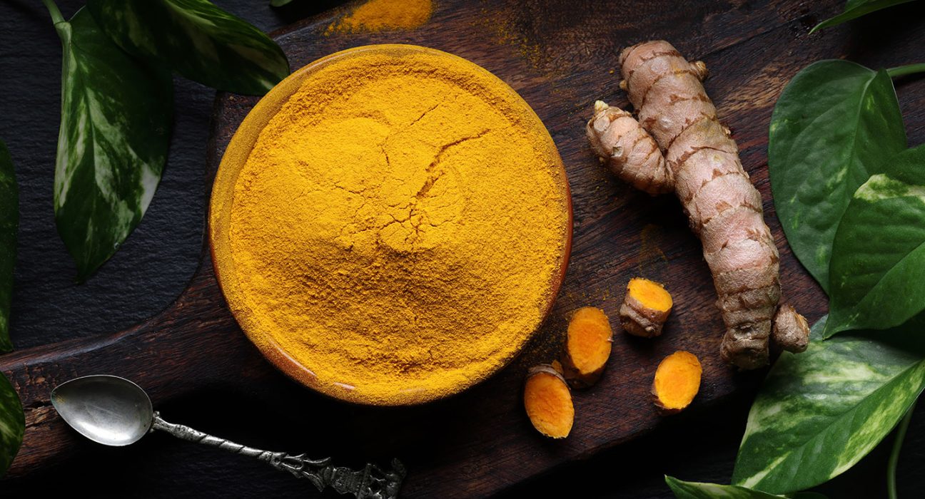 a bowl of bright yellow ground tumeric on a table top next to a tumeric root