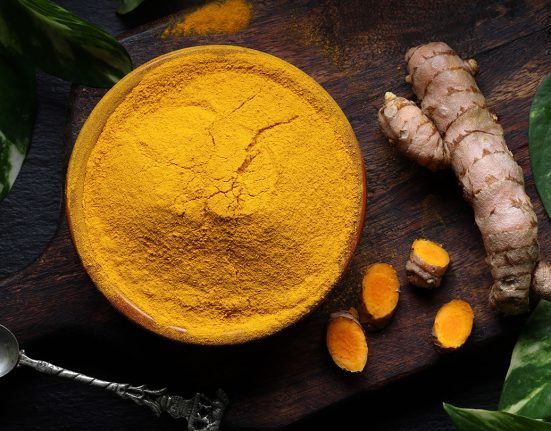 a bowl of bright yellow ground tumeric on a table top next to a tumeric root