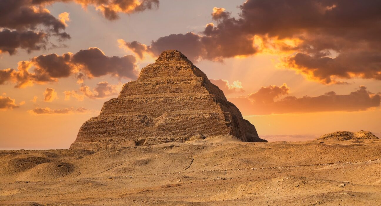 Egyptian King Djoser's pyramid rises above a sandy landscape, while sun-tinged clouds cast a golden glow on the scene.