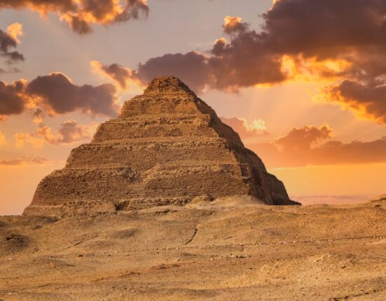 Egyptian King Djoser's pyramid rises above a sandy landscape, while sun-tinged clouds cast a golden glow on the scene.