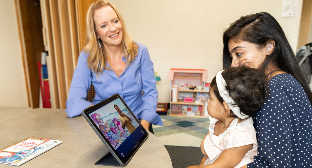 Rain Bosworth smiling and looking at a parent-child pair to her left. She has blonde hair and blue eyes and wearing blue button-up shirt. The parent is looking at an iPad, sitting in front of them on a round table. The iPad is displaying what appears to be a video with a person signing. The parent has black hair and wearing a navy polka dot shirt. The child is sitting on the parent's lap and staring at Bosworth.