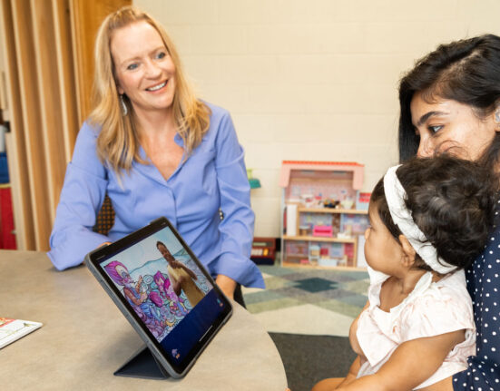 Rain Bosworth smiling and looking at a parent-child pair to her left. She has blonde hair and blue eyes and wearing blue button-up shirt. The parent is looking at an iPad, sitting in front of them on a round table. The iPad is displaying what appears to be a video with a person signing. The parent has black hair and wearing a navy polka dot shirt. The child is sitting on the parent's lap and staring at Bosworth.