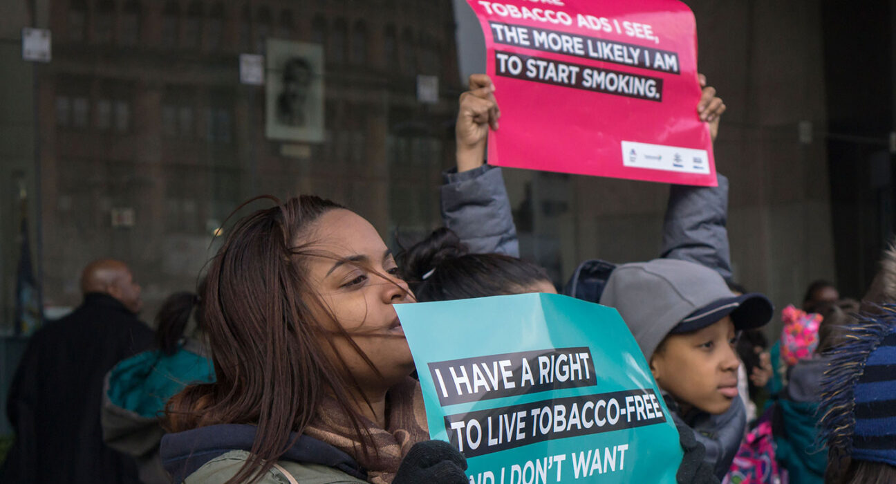 Two young people hold anti-tobacco signs at a a rally. The girl in front holds an teal-colored sign that reads, "I have a right to live tobacco-free." Another person holds a red sign that says, "The more tobacco ads I see, the more likely I am to start smoking."