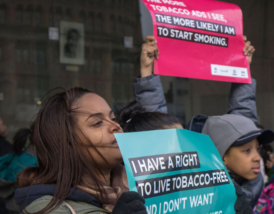 Two young people hold anti-tobacco signs at a a rally. The girl in front holds an teal-colored sign that reads, "I have a right to live tobacco-free." Another person holds a red sign that says, "The more tobacco ads I see, the more likely I am to start smoking."