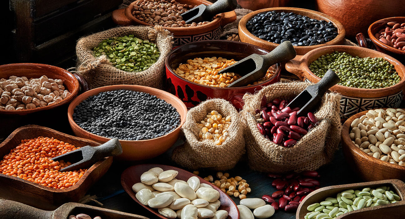 an array of dried beans and legumes arrangesd on a rustic wooden table in bowls and sacks