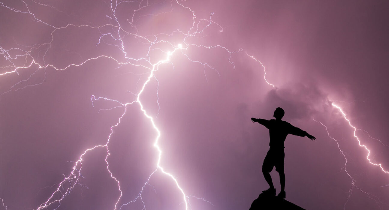 a silhoutte of a person standing on a rocky perch against a purple clouded sky and a lightning strike