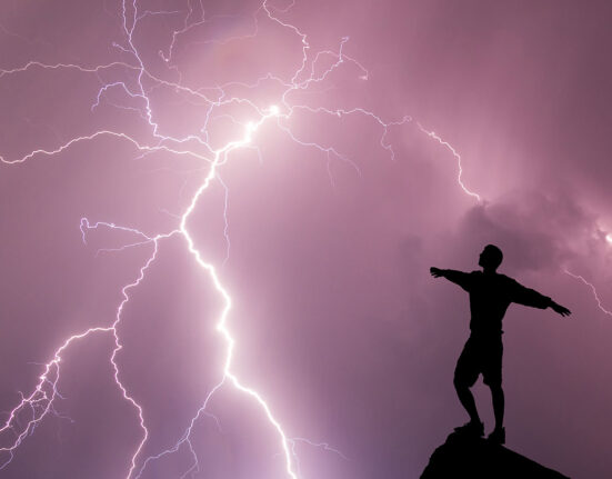 a silhoutte of a person standing on a rocky perch against a purple clouded sky and a lightning strike