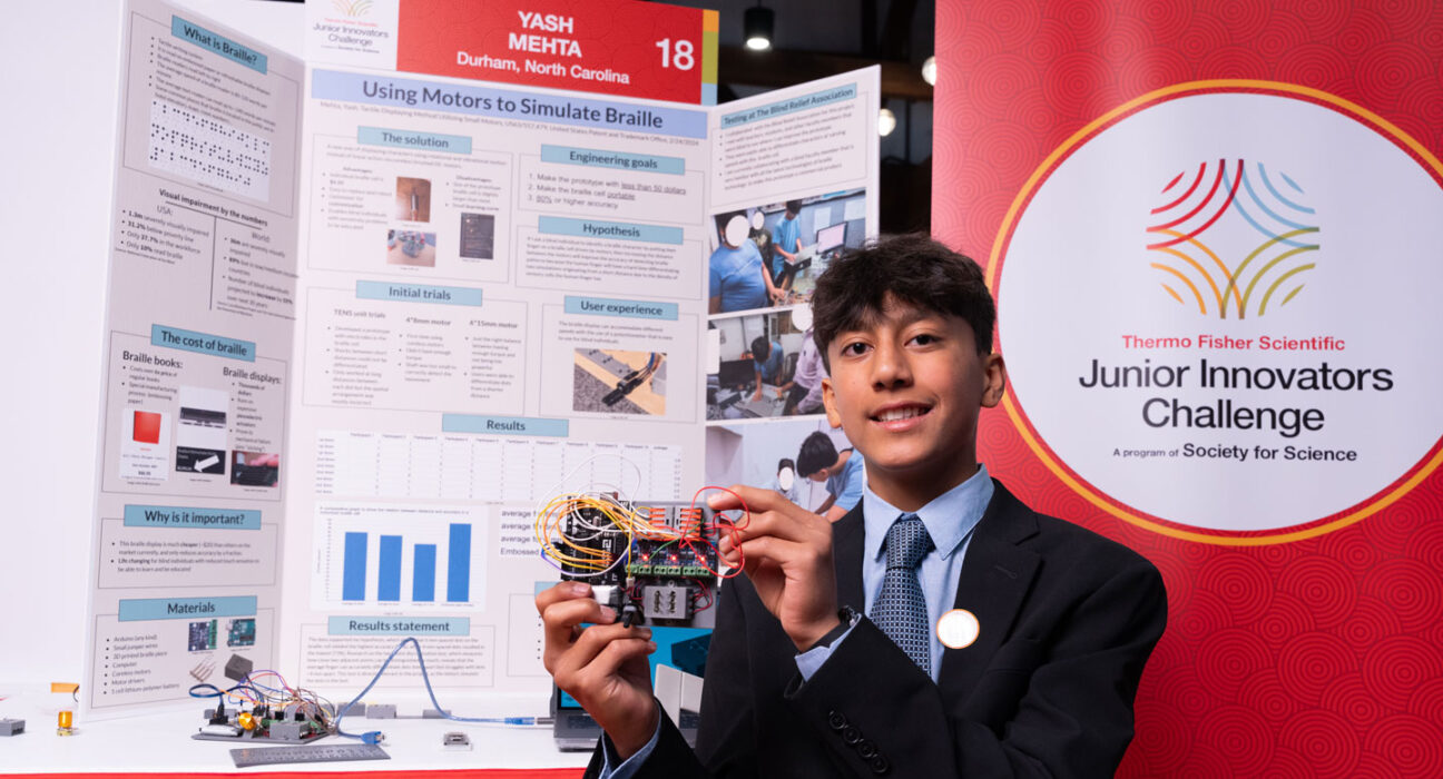 Yash Mehta stands in front of a scientific poster with words and pictures describing his project. The braille reader he holds is an electronic device with several wires and components.