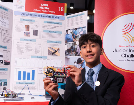 Yash Mehta stands in front of a scientific poster with words and pictures describing his project. The braille reader he holds is an electronic device with several wires and components.