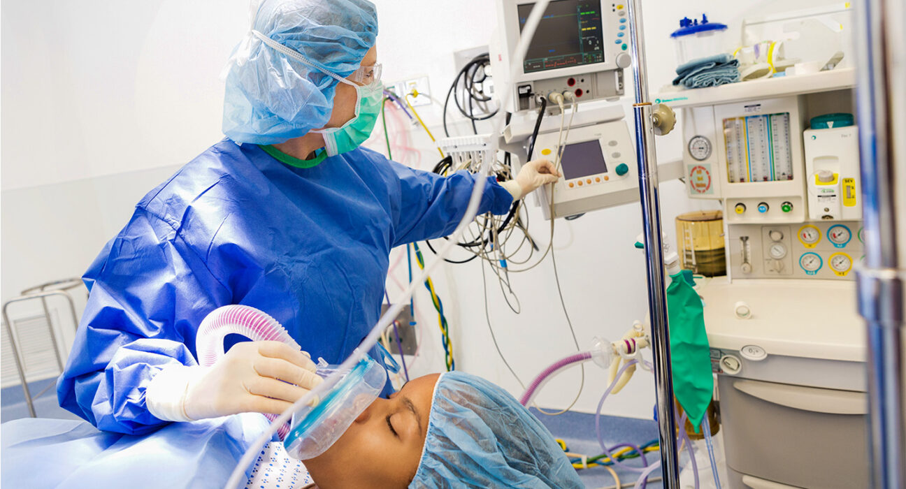 An anesthesiologist in blue scrubs and face mask applies anesthesia to a patient while looking at her monitors.