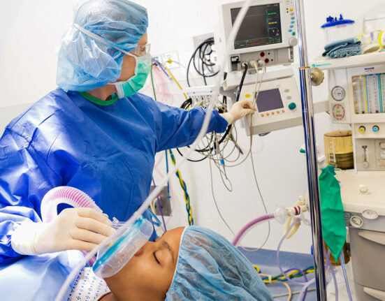 An anesthesiologist in blue scrubs and face mask applies anesthesia to a patient while looking at her monitors.