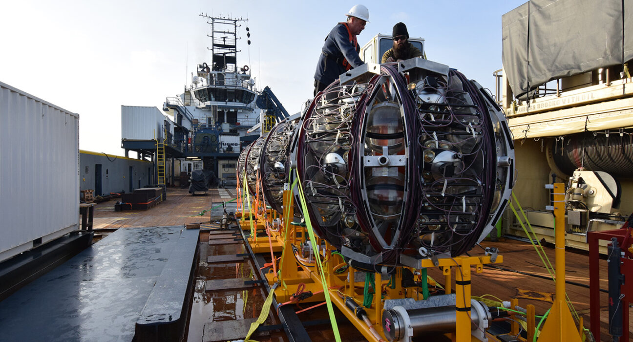 Workers on a ship prepare to launch a giant round piece of equipment off of a bright yellow ramp