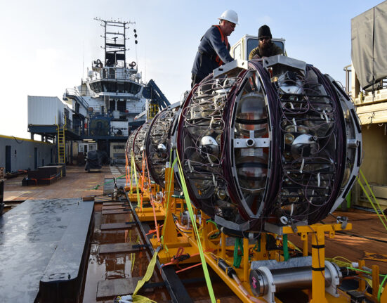 Workers on a ship prepare to launch a giant round piece of equipment off of a bright yellow ramp